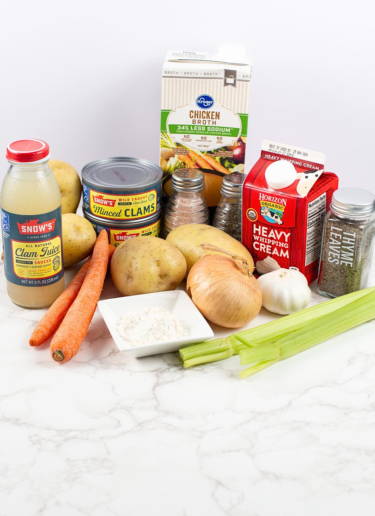A variety of clam chowder ingredients on a white surface, including potatoes, carrots, celery, onion, garlic, canned minced clams, clam juice, chicken broth, heavy cream, flour, dried thyme, salt, and pepper.