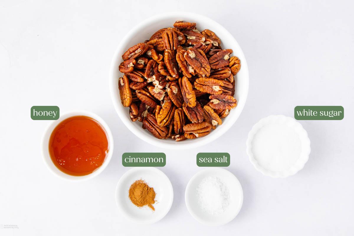A bowl of pecans, and small bowls of honey, white sugar, cinnamon, and sea salt, arranged on a white surface with each ingredient labeled.