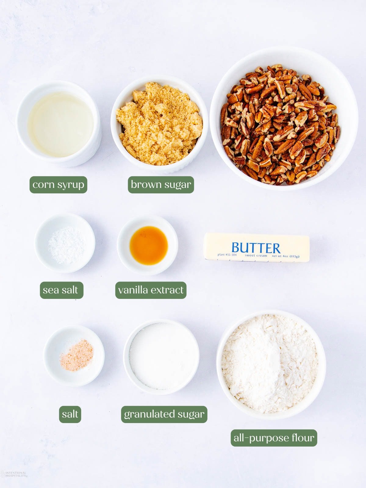 Overhead view of various baking ingredients in bowls, labeled as: corn syrup, brown sugar, pecans, sea salt, vanilla extract, butter, salt, granulated sugar, all-purpose flour. Items are arranged on a white surface.