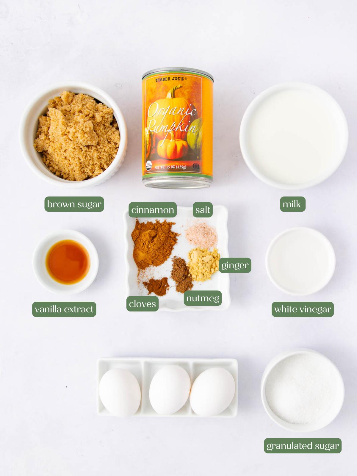 A flat lay of ingredients for baking, including canned pumpkin, milk, brown sugar, vanilla extract, granulated sugar, eggs, white vinegar, and small bowls of spices: cinnamon, salt, ginger, cloves, and nutmeg.