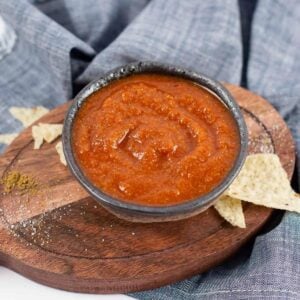 A small black bowl filled with red salsa sits on a wooden serving board, surrounded by a few tortilla chips, with a gray cloth in the background.