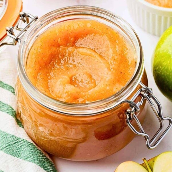 A glass jar filled with smooth, light brown applesauce sits on a white surface next to a green apple, apple slices, and a green-striped cloth. The jar has a metal latch and lid.