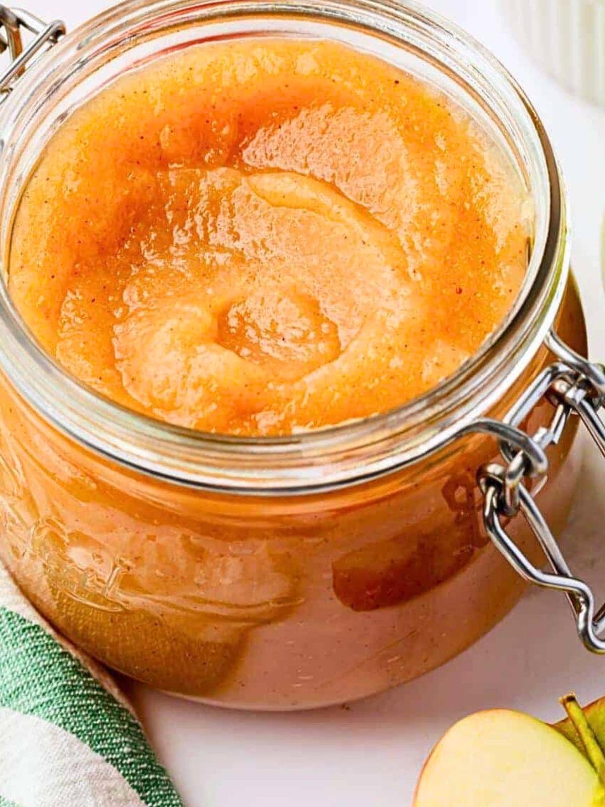A close-up of a glass jar filled with smooth homemade applesauce, with a metal clasp lid. A green and white towel and a slice of apple are partially visible beside the jar.