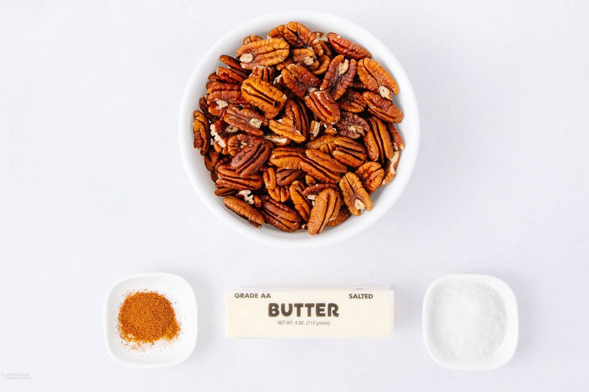 A bowl of pecan halves, a stick of salted butter, a small dish of salt, and a small dish of orange-brown seasoning are arranged on a white background.