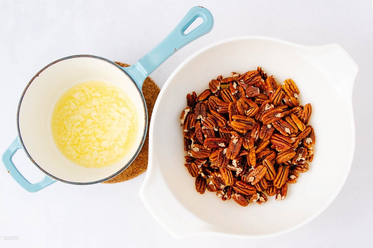 A white bowl filled with pecan halves sits next to a small saucepan containing melted butter on a cork trivet, all on a white surface.