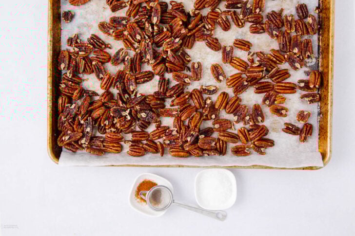 A baking sheet lined with parchment paper holds roasted pecans. In front are a small bowl of salt, a bowl of mixed spices, and a measuring spoon.