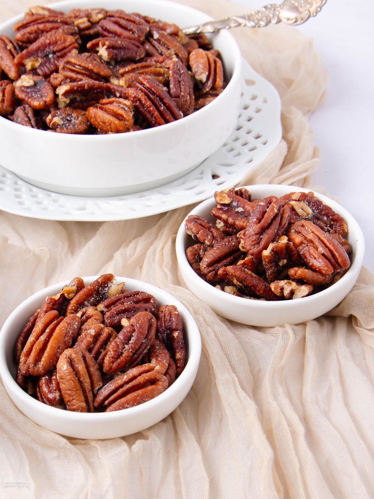Three white bowls filled with glazed pecans are arranged on a light beige cloth. The largest bowl is on a decorative plate, while two smaller bowls are placed nearby. A silver spoon rests in the largest bowl.