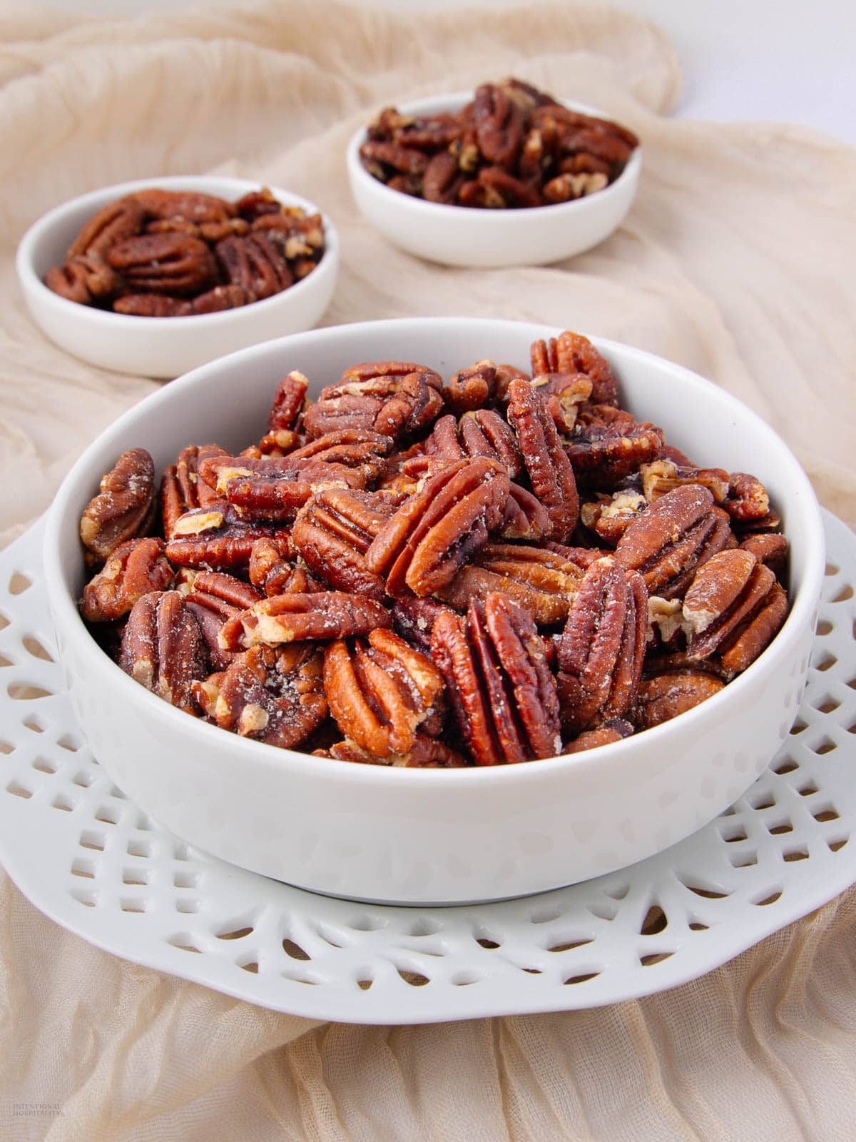 A close-up of a white bowl filled with pecans, with two smaller bowls of pecans in the background, all placed on a white plate and beige fabric.