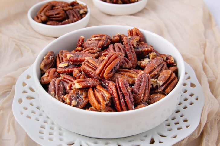 A white bowl filled with glazed pecans sits on a white decorative plate, with two smaller bowls of pecans in the background on a light-colored fabric.