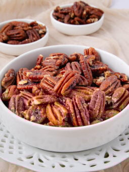 A white bowl filled with glazed pecans sits on a decorative white plate, with two smaller bowls of pecans in the background, all placed on a beige fabric.