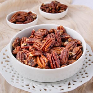 A white bowl filled with glazed pecans sits on a decorative white plate, with two smaller bowls of pecans in the background, all placed on a beige fabric.