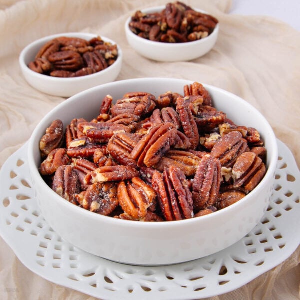 A white bowl filled with glazed pecans sits on a decorative white plate, with two smaller bowls of pecans in the background, all placed on a beige fabric.