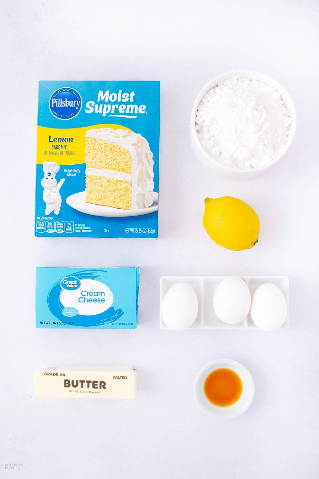 Top-down view of ingredients for baking: a box of lemon cake mix, powdered sugar, a lemon, a block of cream cheese, three eggs in a tray, a stick of butter, and a small bowl of vanilla extract.
