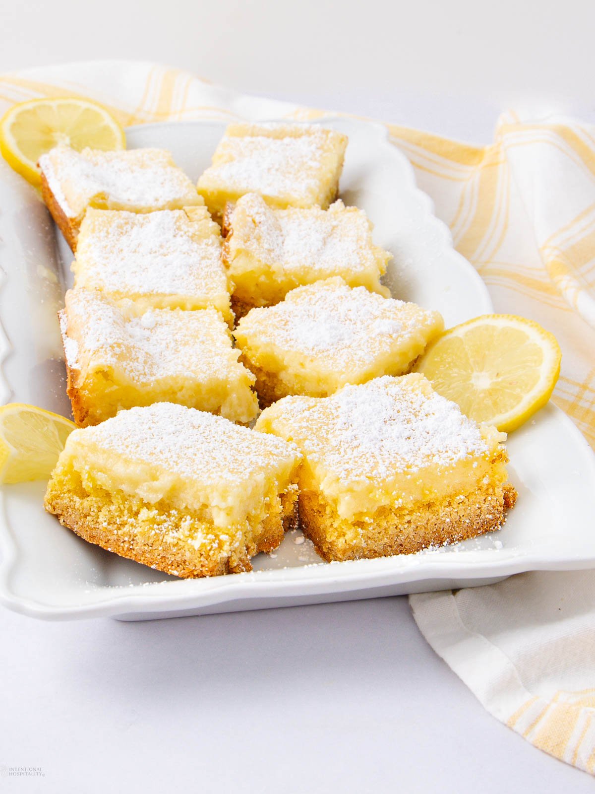 A white platter displays several lemon bars topped with powdered sugar, arranged in rows. Lemon slices are placed as garnish, and a yellow-and-white striped cloth is partially visible beside the platter.