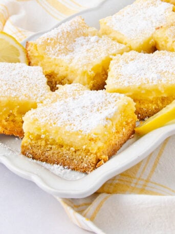 Squares of lemon bars with a golden crust and powdered sugar on top are arranged on a white plate, with lemon slices and a yellow plaid napkin nearby.