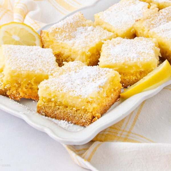 Squares of lemon bars with a golden crust and powdered sugar on top are arranged on a white plate, with lemon slices and a yellow plaid napkin nearby.