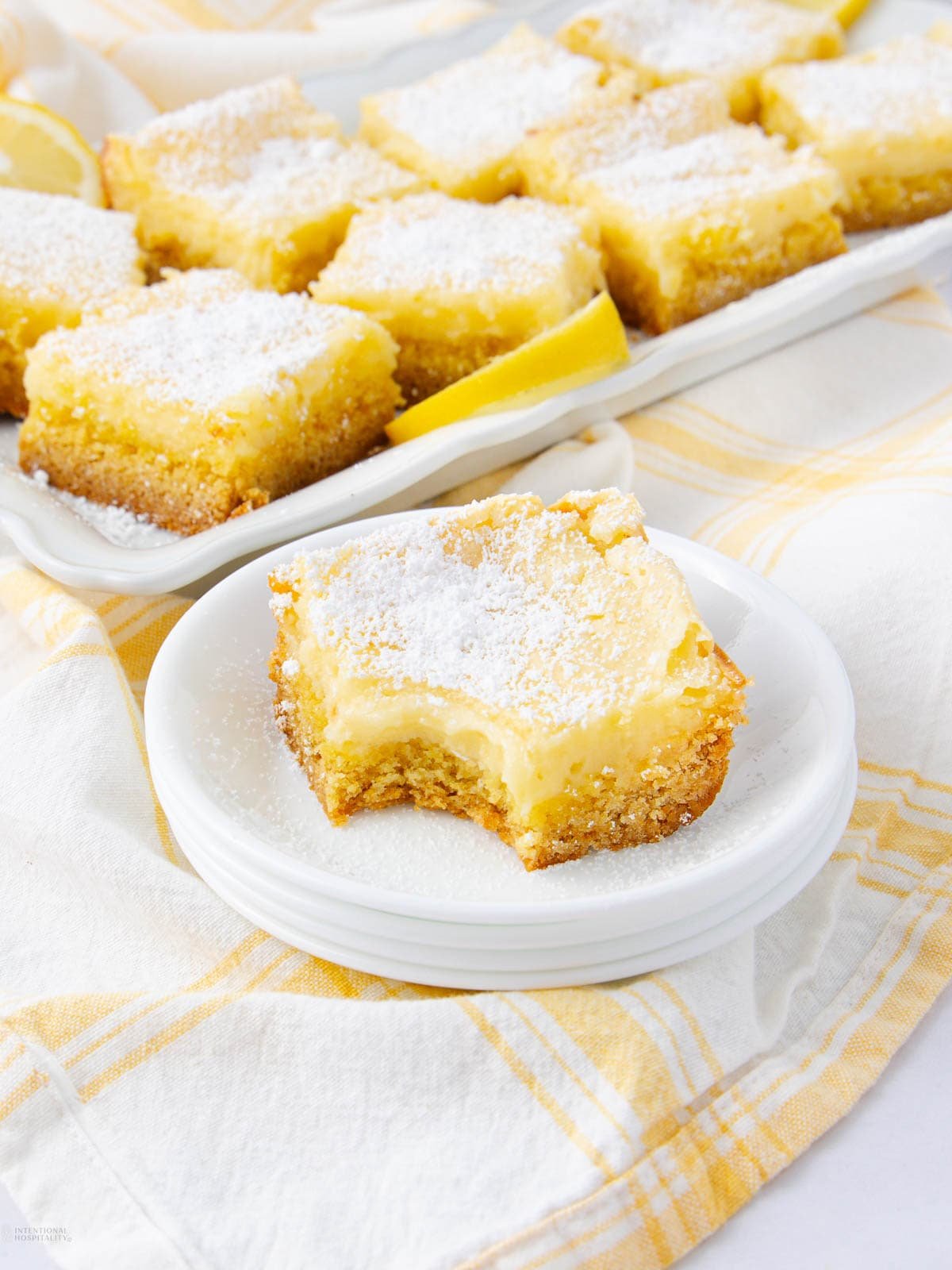 A plate with a lemon bar that has a bite taken out of it, dusted with powdered sugar. More lemon bars are arranged on a tray in the background, all on a yellow and white checkered cloth.