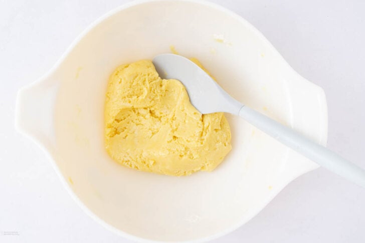 A white mixing bowl containing a ball of yellow dough is being stirred with a white spatula against a white background.