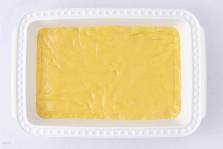 A white rectangular baking dish filled with an even, unbaked layer of yellow dough, smooth and pressed flat across the bottom. The dish sits on a white surface.