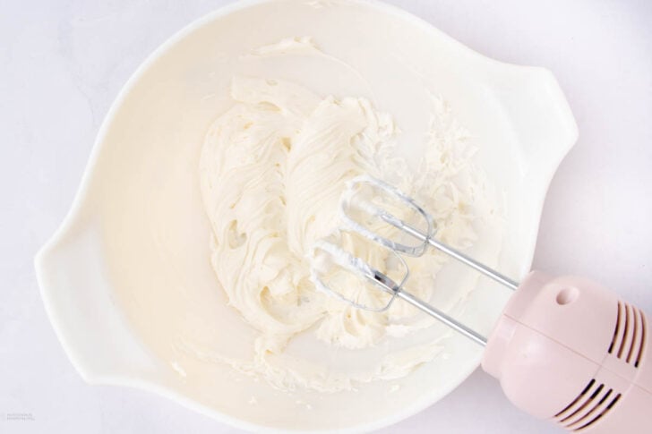 A white mixing bowl containing whipped cream cheese being mixed with a pink electric hand mixer, resting on a white surface.