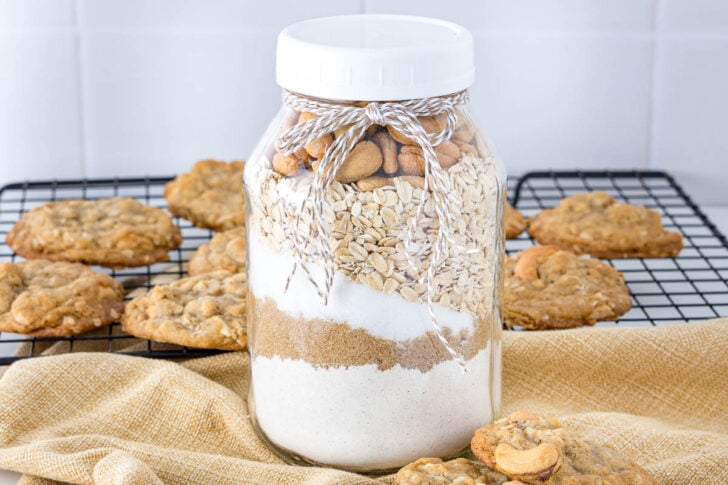 A glass jar filled with layers of flour, sugar, oats, and nuts, tied with twine, sits in front of a cooling rack with oatmeal cookies on a light cloth.