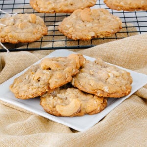 A plate with four cashew cookies sits on a beige cloth, with more cookies cooling on a wire rack in the background. The cookies are golden brown with visible cashew pieces.