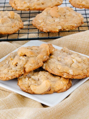 A plate with four cashew cookies sits on a beige cloth, with more cookies cooling on a wire rack in the background. The cookies are golden brown with visible cashew pieces.