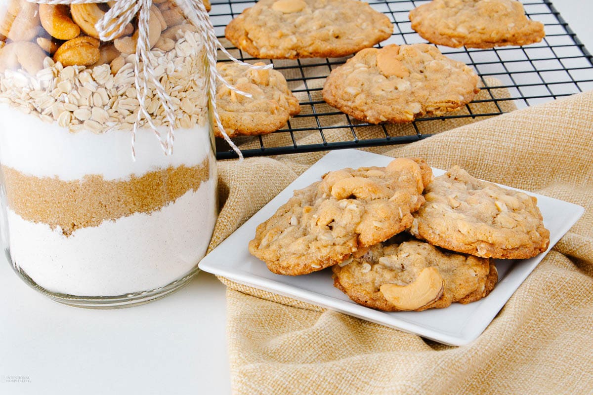 A plate of homemade cookies sits beside a jar filled with layered cookie ingredients and a cooling rack with more cookies in the background. A beige cloth is placed under the plate.