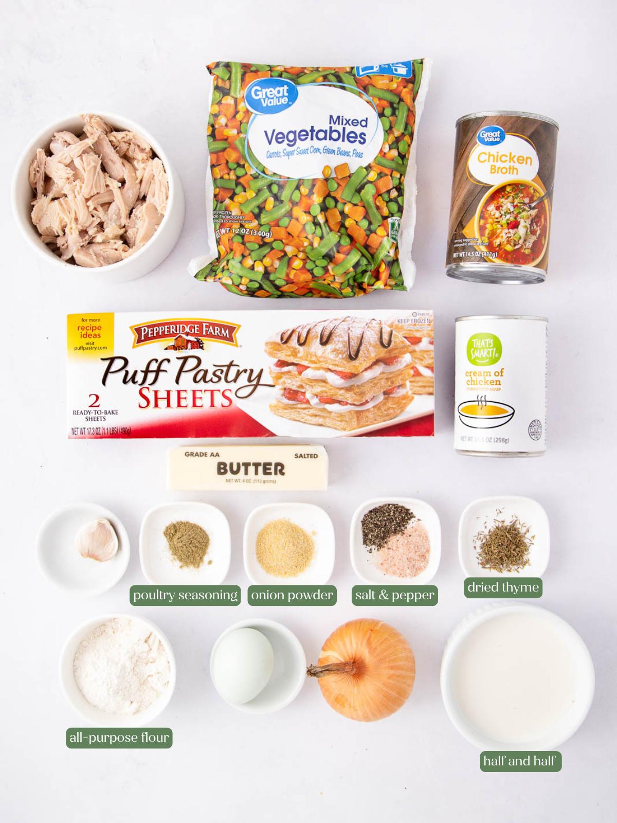 Ingredients for chicken pot pie are arranged on a white surface, including mixed vegetables, canned chicken broth, puff pastry, cooked chicken, cream of chicken soup, seasonings, flour, butter, half and half, and an onion.