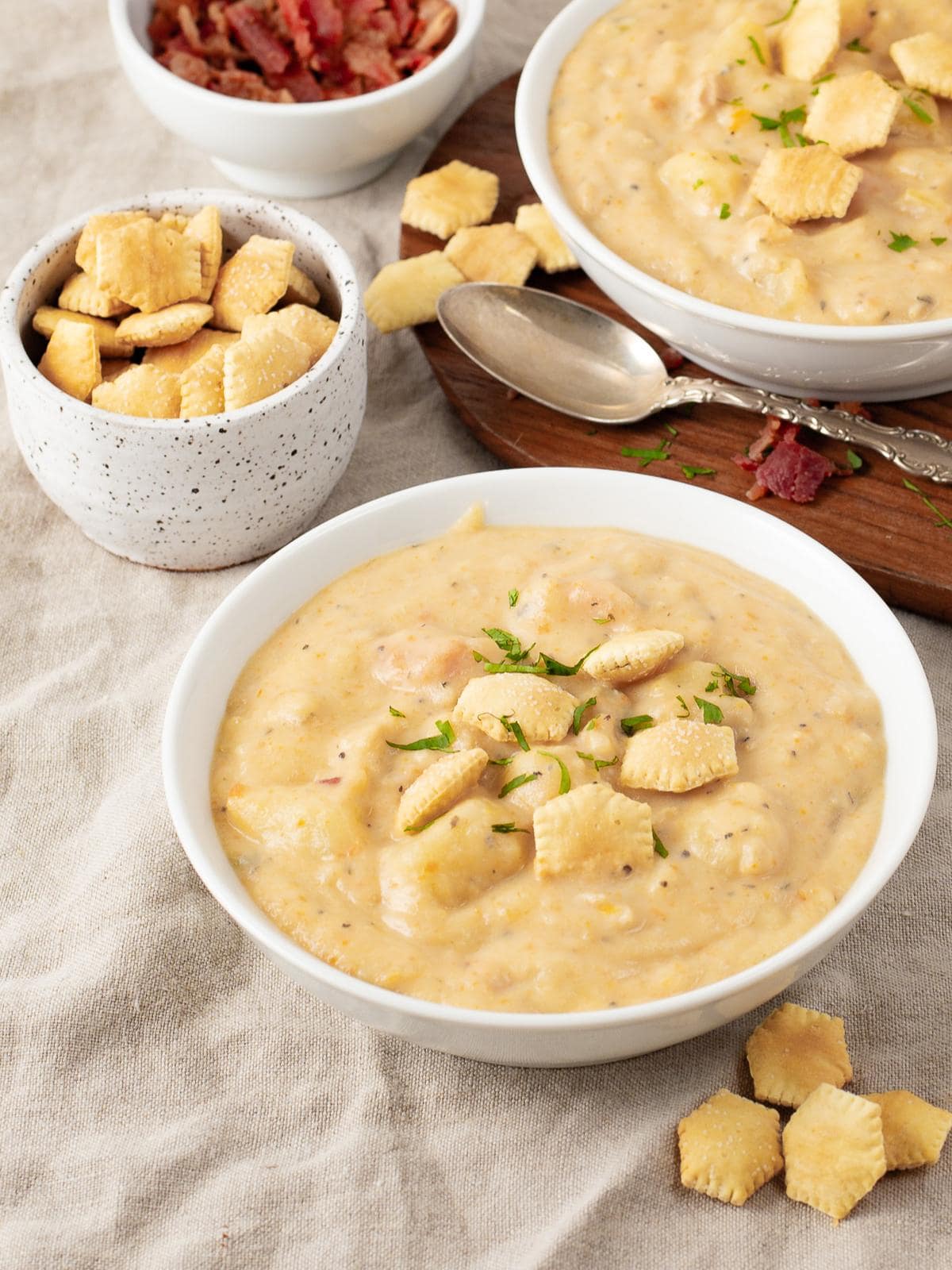 A bowl of creamy soup topped with oyster crackers and fresh herbs sits on a cloth next to a bowl of crackers, a bowl of crumbled bacon, and a spoon. Another bowl of soup is partially visible in the background.