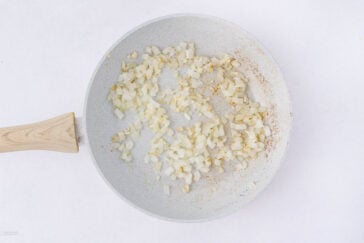 A white frying pan with light wood handle contains diced onions being sautéed, starting to turn translucent and slightly golden, on a white background.