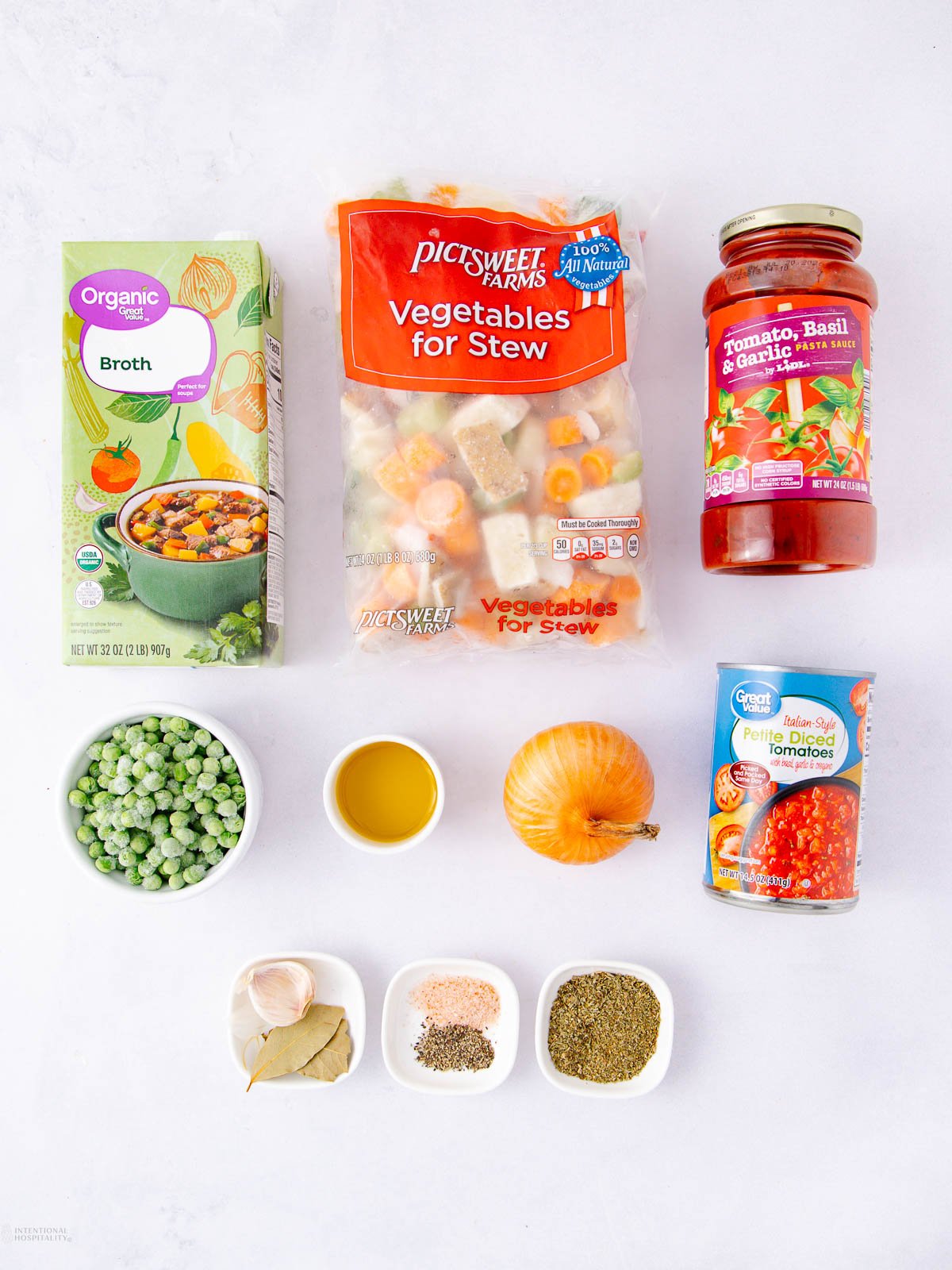 Assorted ingredients for a vegetable stew are arranged on a white surface, including vegetable broth, mixed stew vegetables, pasta sauce, frozen peas, olive oil, an onion, garlic, seasonings, and a can of diced tomatoes.