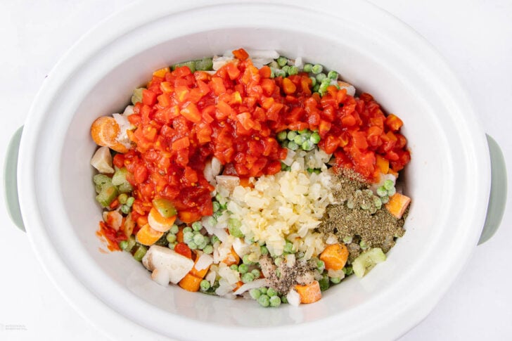 A slow cooker filled with chopped carrots, peas, celery, onions, minced garlic, diced tomatoes, and various dried herbs, ready to be cooked.