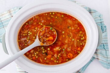 A white slow cooker filled with vegetable soup; visible ingredients include carrots, peas, corn, and tomatoes. A ladle rests inside the pot. The slow cooker sits on a blue and white plaid cloth.