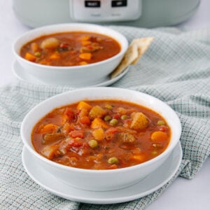 Two white bowls of hearty vegetable soup with visible carrots, potatoes, peas, and tomatoes sit on matching plates atop a light plaid cloth, with a slow cooker in the background and crackers on the side.