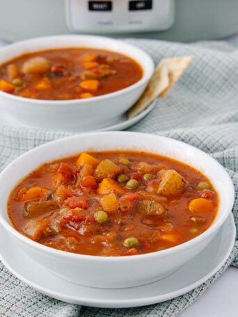 Two white bowls of hearty vegetable soup with visible carrots, potatoes, peas, and tomatoes sit on matching plates atop a light plaid cloth, with a slow cooker in the background and crackers on the side.