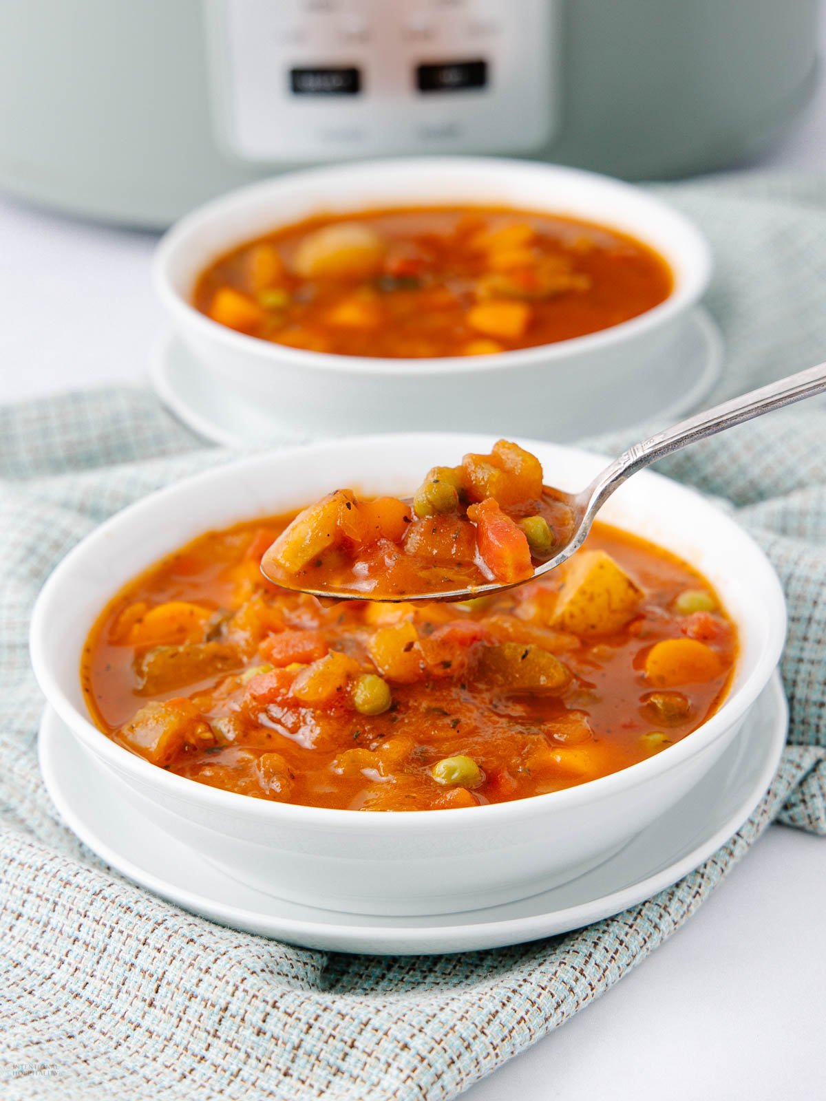 Two white bowls of chunky vegetable soup with carrots, peas, and potatoes sit on saucers. A spoon holds a scoop of the soup above one bowl. A textured cloth and a slow cooker are in the background.