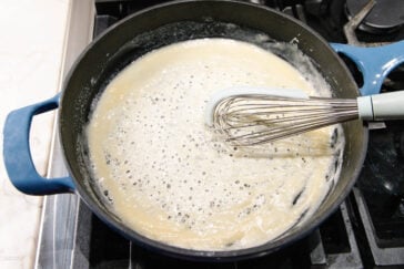 A blue pot sits on a stove with a light-colored roux being whisked; bubbles are forming on the surface as the mixture cooks. A metal whisk with a light blue handle rests inside the pot.