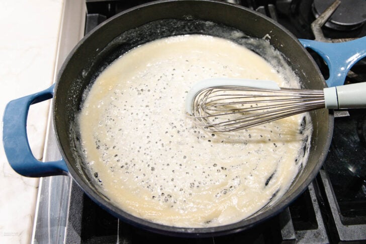 A blue pot sits on a stove with a light-colored roux being whisked; bubbles are forming on the surface as the mixture cooks. A metal whisk with a light blue handle rests inside the pot.