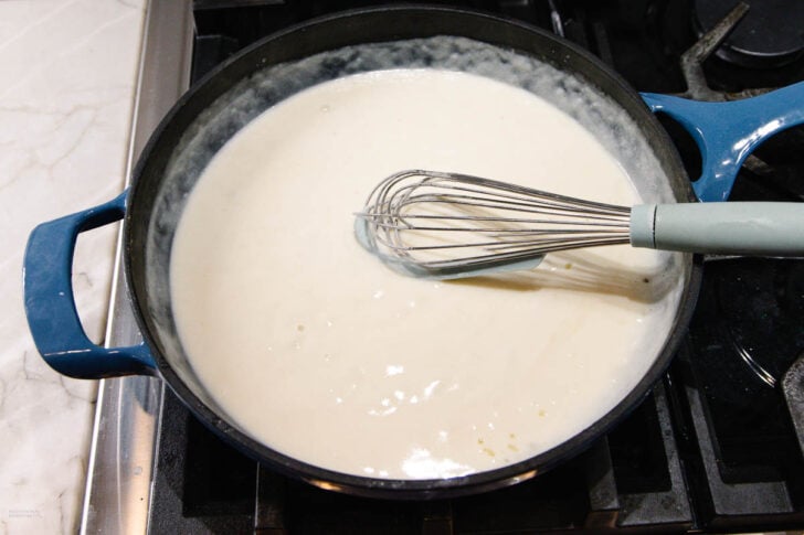 A whisk stirring creamy white sauce in a blue pot on a stovetop, with part of the marble countertop visible beside the stove.