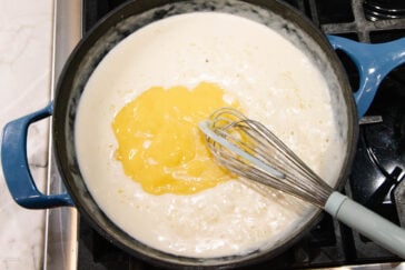 A whisk mixes melted butter into a creamy white sauce in a blue pan on a stovetop.