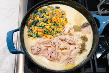 A blue pot on a stovetop filled with creamy sauce, shredded chicken, frozen mixed vegetables, and assorted dried herbs and seasonings, all ready to be mixed together.