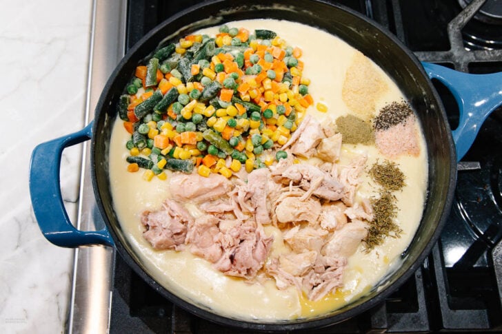 A blue pot on a stovetop filled with creamy sauce, shredded chicken, frozen mixed vegetables, and assorted dried herbs and seasonings, all ready to be mixed together.