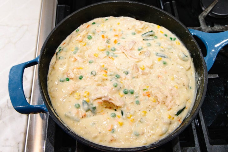 A blue pot on a stovetop is filled with a thick, creamy mixture containing shredded chicken, peas, corn, carrots, and green beans. The dish appears to be in the process of being cooked.