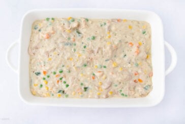 A white baking dish filled with a creamy mixture containing chunks of chicken, green peas, corn, carrots, and other vegetables, on a white background.
