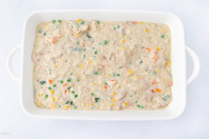 A white baking dish filled with a creamy mixture containing chunks of chicken, green peas, corn, carrots, and other vegetables, on a white background.