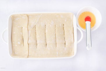 A white baking dish with puff pastry sheets laid out and sliced into strips. Next to it is a bowl of beaten egg and a pastry brush. All items are on a white surface.
