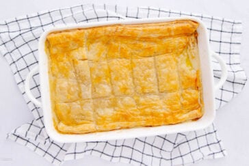 A rectangular white baking dish with a golden, flaky puff pastry pie sits on a white and black checkered kitchen towel. The pastry is evenly baked and lightly scored into sections.