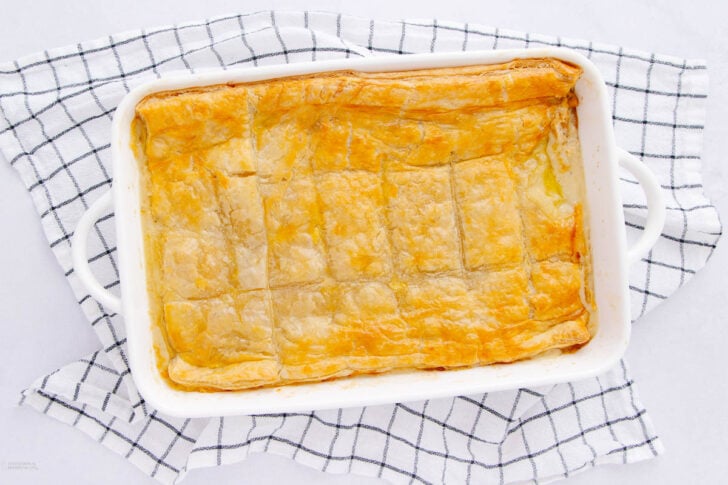 A rectangular white baking dish with a golden, flaky puff pastry pie sits on a white and black checkered kitchen towel. The pastry is evenly baked and lightly scored into sections.