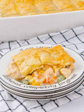 A slice of creamy chicken pot pie with a golden crust on a plate, showing visible vegetables, set on a stack of plates with text, beside a larger dish of pot pie on a checkered cloth.